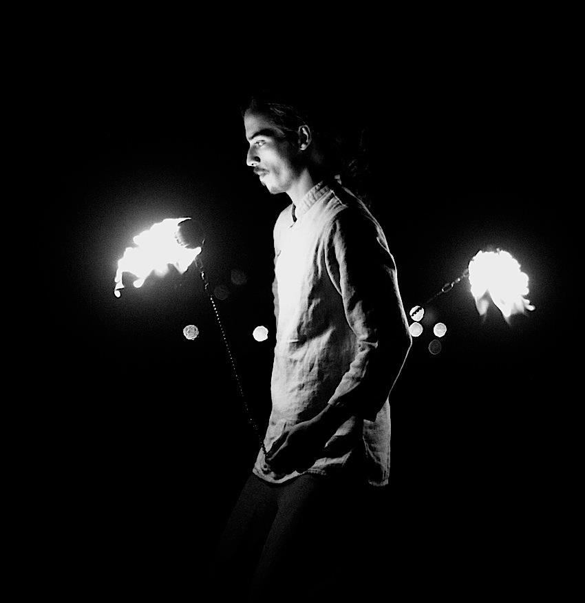 Black and white, high-contrast photo of a young man spinning fire poi. His expression is calm and void-like.