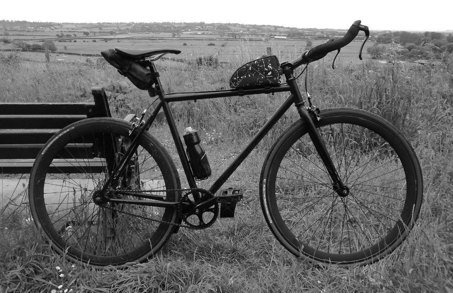 A black fixed gear bicycle beside a bench, with rolling green fields in the background.
