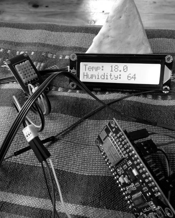 Black and white photo in portrait orientation showing electronics on a striped table-cloth. A NodeMCU dev board appears in the lower-right, DHT11 sensor in the top-left, and an LCD in the top-right. Text on the display reads: temp: 18.0 and humidity: 64. The display is propped-up by a white pyramid (mycelium) and a wooden floor is just visible in the background.