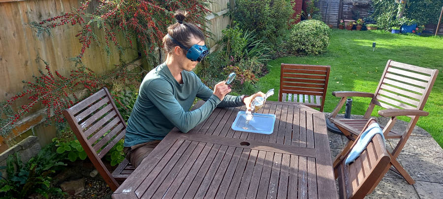 The artist etching a glyph with a magnifying glass. He is seated at a wooden table and wearing welding goggles. A garden scene is visible in the background.